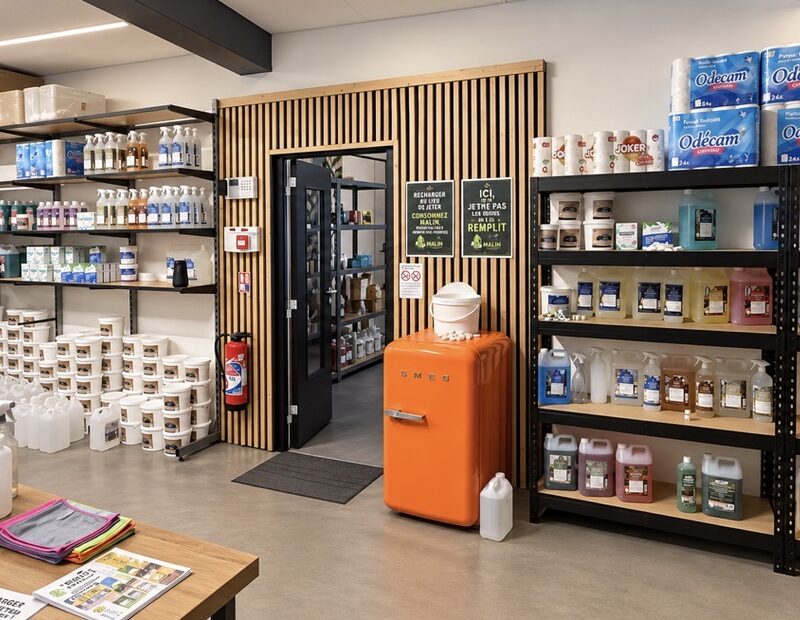 Intérieur de l’entrepôt du Bidon Malin : produits d’entretien en recharge, contenants réutilisables et affiche « Recharger au lieu de jeter »